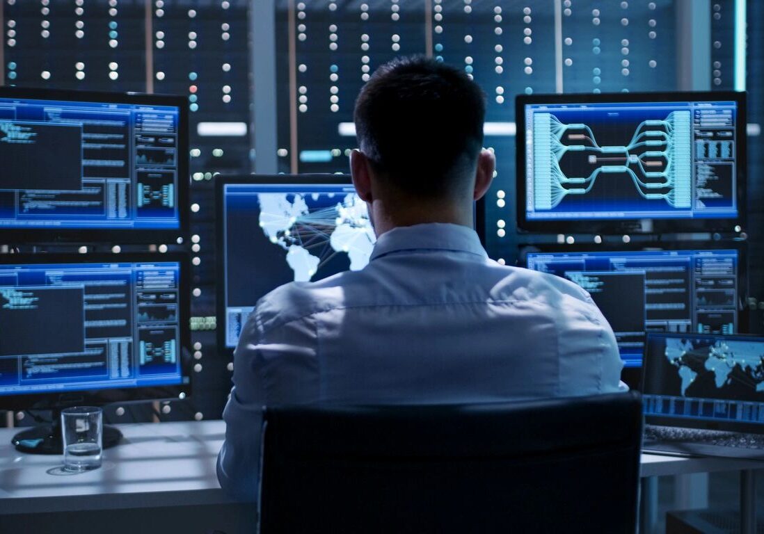 Man analyzing multiple digital data screens in a tech control room.
